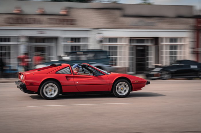 A red Ferrari 308 driving past a row of storefronts, panned so the background blurs.