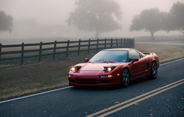 A red Acura NSX with its headlights on, on a fog-covered country road at dawn.