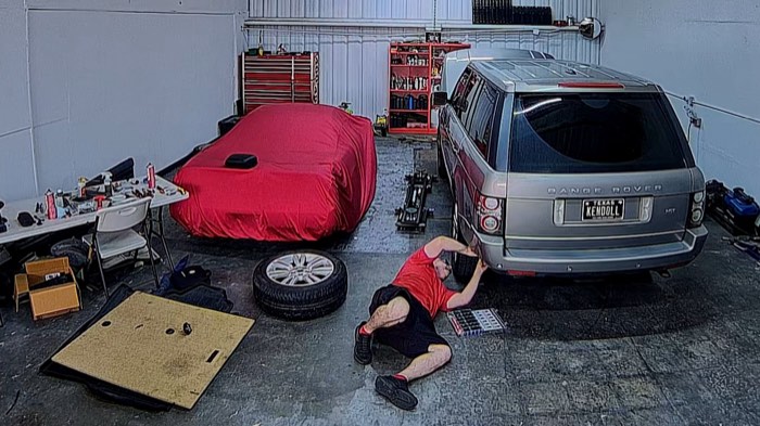 Robert Cope on the shop floor, working behind the rear wheel of a silver Range Rover with the wheel removed; a red car cover and tool chests fill the background.